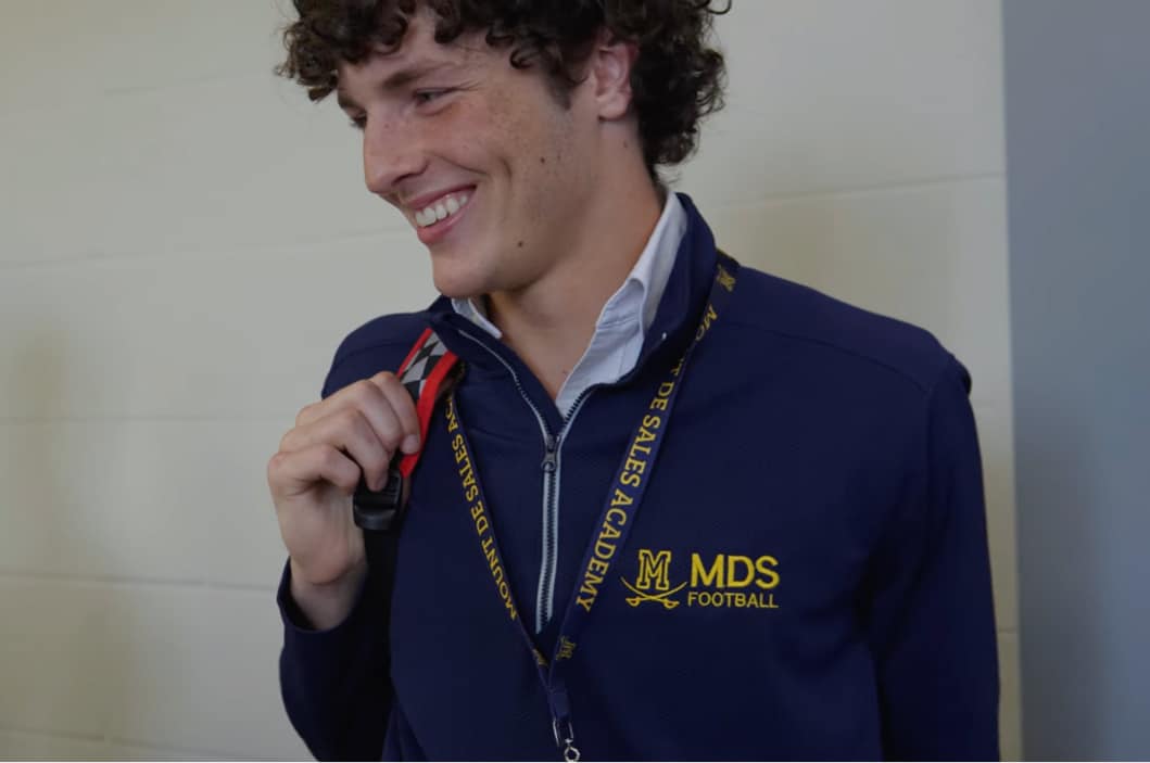 A young man with curly hair smiles while wearing a navy MDS Football jacket and matching lanyard, holding a red strap indoors.