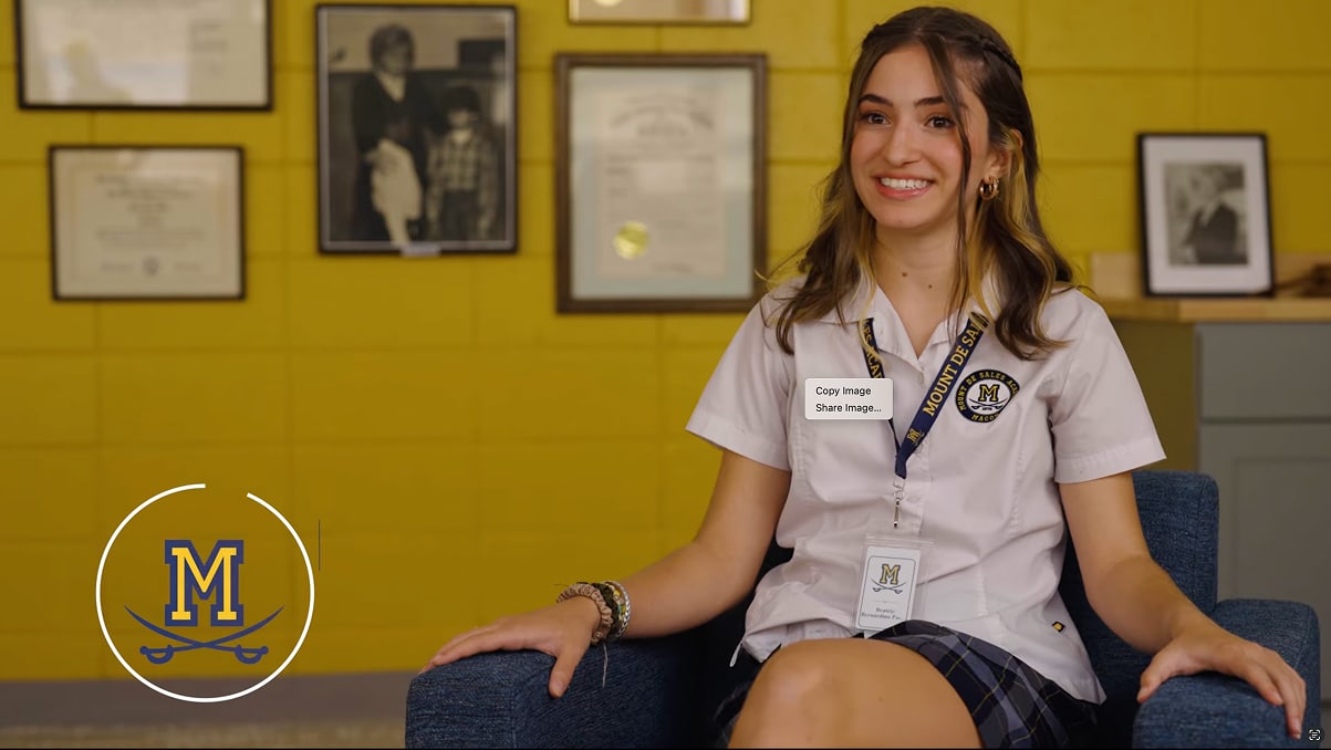 A smiling young woman in a school uniform sits on a chair. She has long brown hair, a badge, plaid skirt; yellow wall behind her.